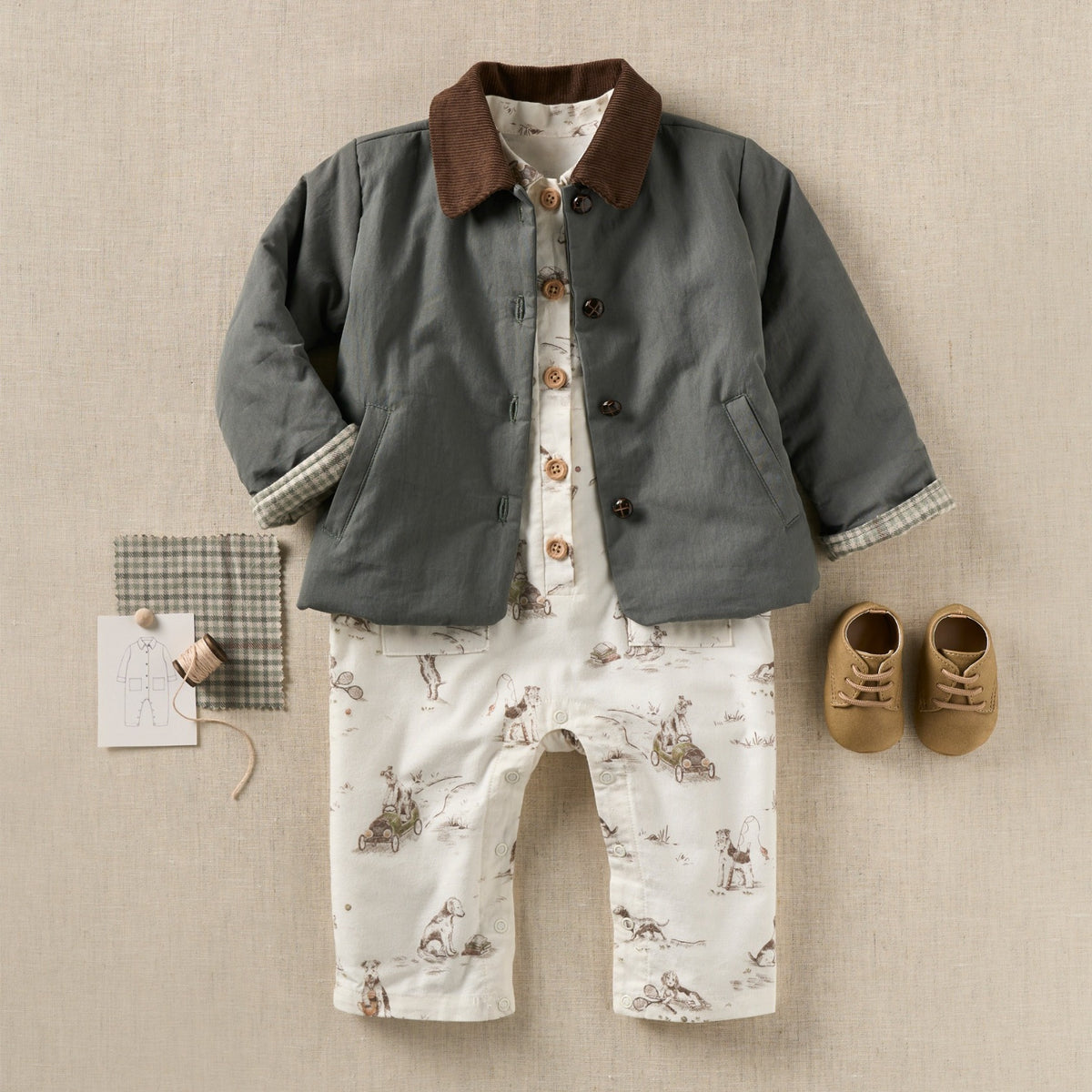 zoomed in A baby boy's two-piece outfit set featuring a dark gray quilted jacket with a brown corduroy collar and white pants with a brown animal print, styled with a pair of tan baby shoes and a small gingham accessory on a textured beige background.