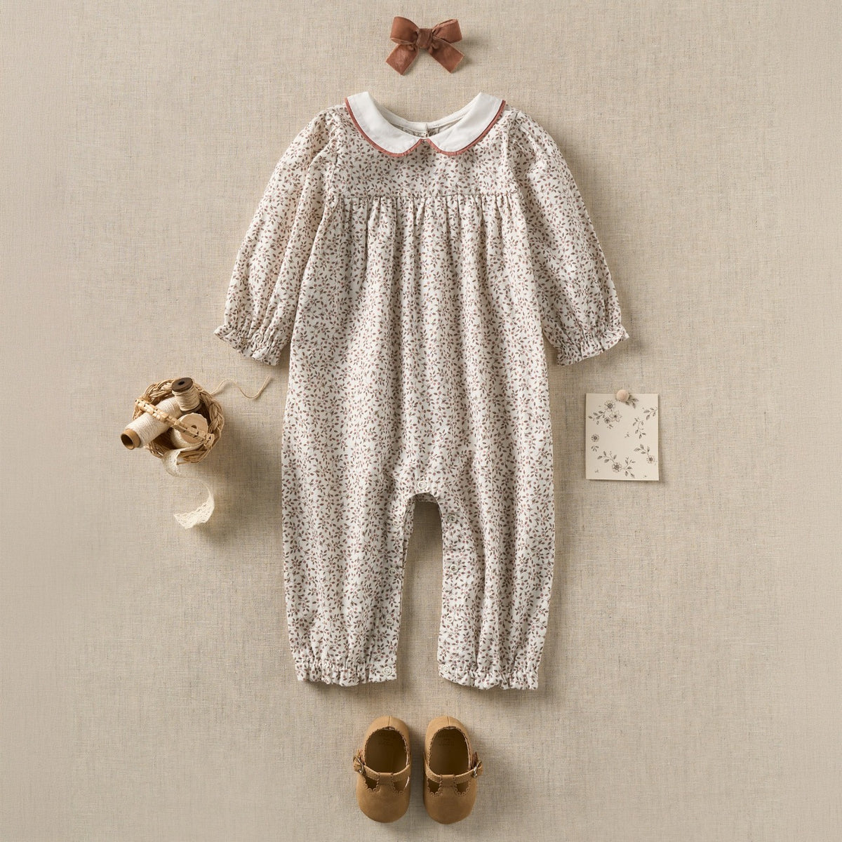 A baby girl's long-sleeve floral romper in a light beige and white ditsy print, styled in a flat-lay arrangement with a matching brown bow, a small card, baby shoes, and a tiny hat on a textured beige background.