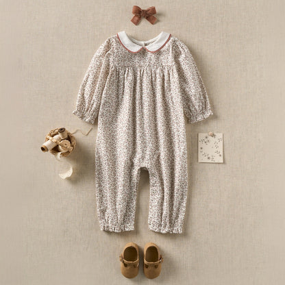 A baby girl's long-sleeve floral romper in a light beige and white ditsy print, styled in a flat-lay arrangement with a matching brown bow, a small card, baby shoes, and a tiny hat on a textured beige background.