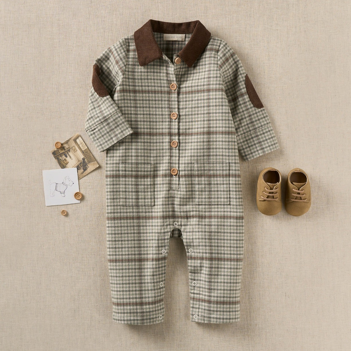 zoomed in A baby long-sleeve romper laid flat on a beige textured background, featuring a muted green and brown plaid pattern, a solid dark brown collar, brown elbow patches, and wooden buttons. A pair of tan baby shoes and small decorative cards are placed beside it.