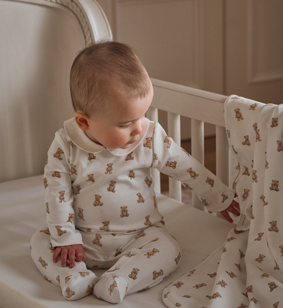 A baby sitting in a white crib wearing a cream footed one-piece pajama with a brown teddy bear pattern, while touching a matching teddy bear print blanket.